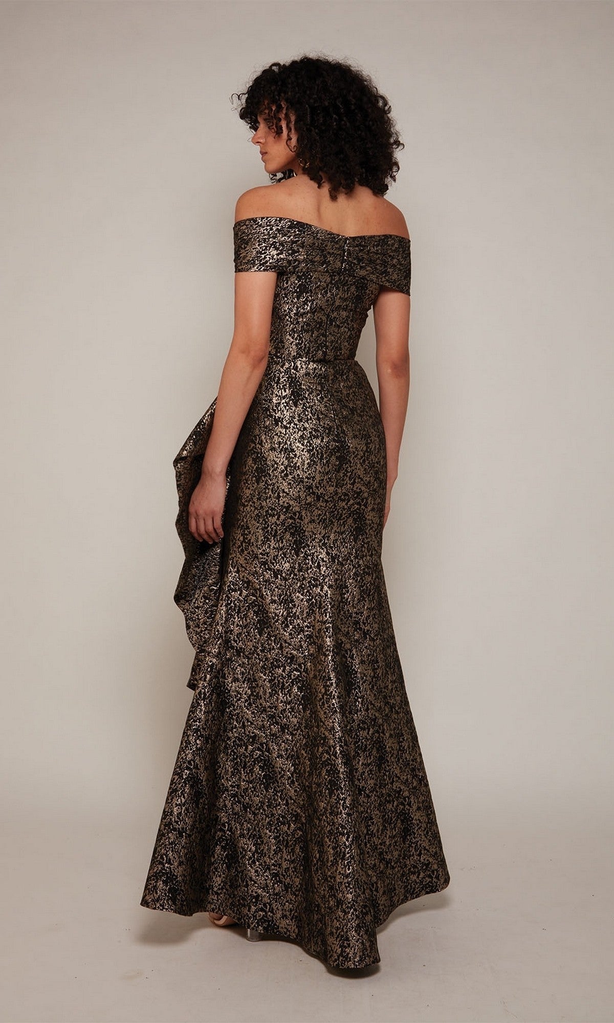 A woman faces away, wearing the Alyce 27667—a metallic jacquard, off-the-shoulder, floor-length gown with a floral pattern and side drape—perfect for formal occasions or as a mother-of-the-bride dress.